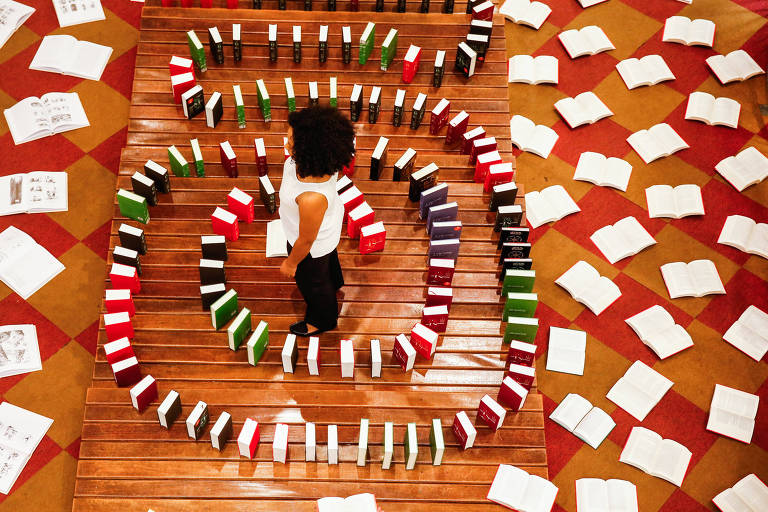 Mulher em pé em meio a um caracol de livros, organizados como uma espécie de "efeito dominó", sem aparecer as capas