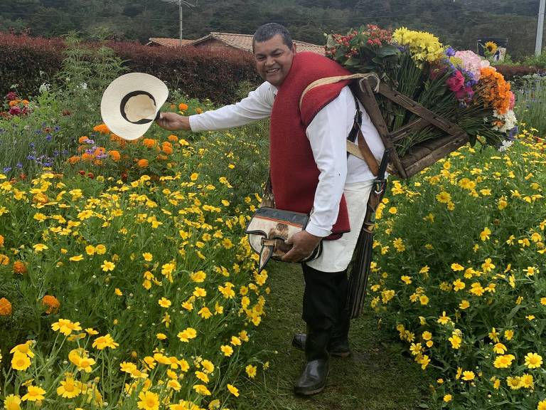 Silletero com silleta tradicional de flores tira chapéu e sorri para foto entre flores na fazenda