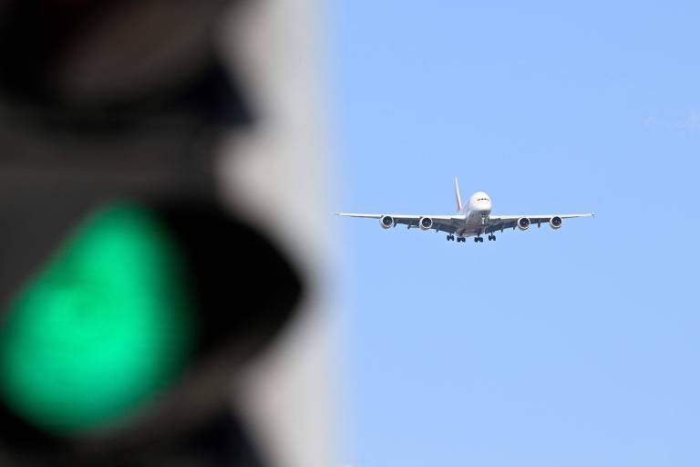 Imagem mostra avião voando sob céu ensolarado. No canto direito da foto, em primeiro plano, há um semáforo no sinal verde.