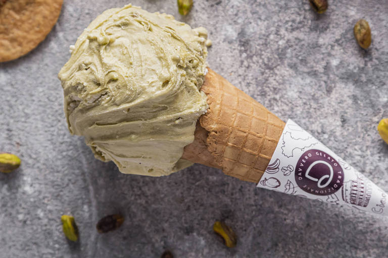 Na foto, uma casquinha de sorvete com uma bola de pistache aparece apoiada em um fundo acinzentando com pistaches