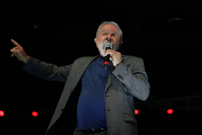 Vestido com paletó cinza e blusa azul diante de um fundo preto com luzes vermelhas, Lula, um homem branco de barba e cabelo da mesma cor, discursa com o microfone na mão esquerda e outro braço estendido com o delo apontado