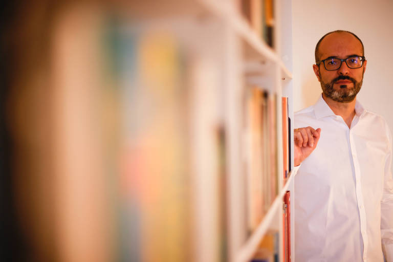 Homem branco com barba escura e calvície pronunciada. Usa óculos e veste camisa branca. Está em pé apoiado numa estante de livros.