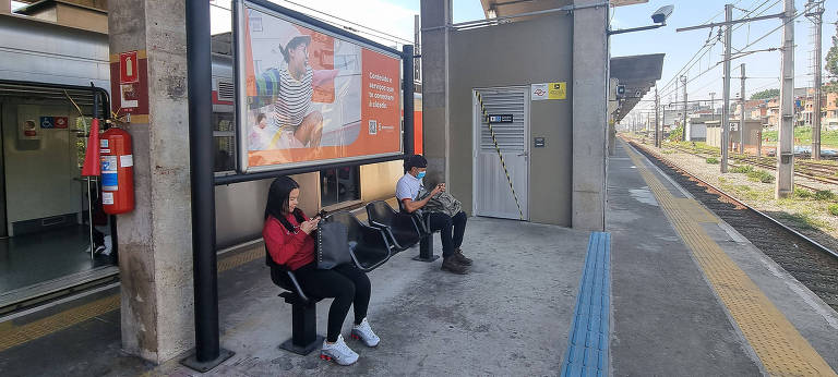 Imagem mostra trem da CPTM parado em estação. Duas pessoas estão sentadas em um banco.