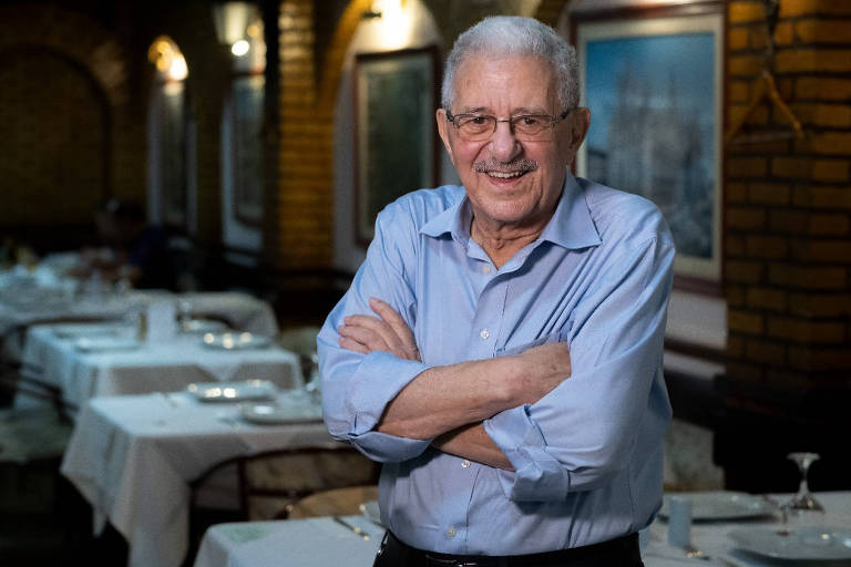 Foto de Simon Pitel, um homem branco e idoso, de cabelos brancos. Ele está de camisa azul, braços cruzados e sorri para câmera. Ao fundo, mesas postas dentro de restaurante.