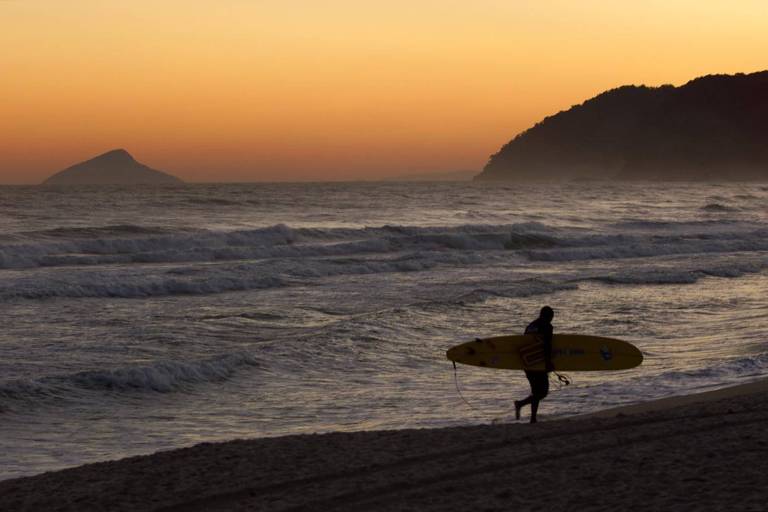 Pessoa anda com uma prancha de surfe na praia num pôr do sol