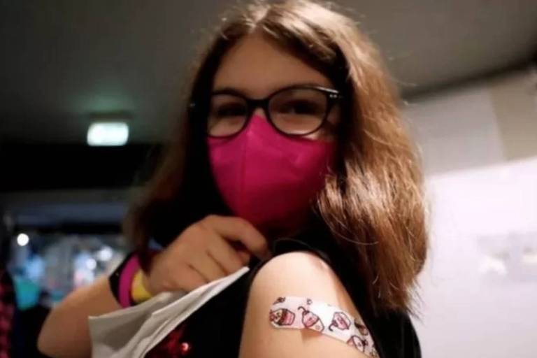 Foto de menina de cabelos lisos, usando óculos e máscara, mostrando o band-aid no braço onde a vacina foi aplicada.