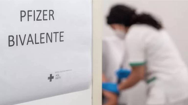 Foto mostrando, ao fundo, uma enfermeira aplicando a vacina em um paciente. Na entrada da sala, a foto foca na placa da porta, escrito "Pfizer bivalente".