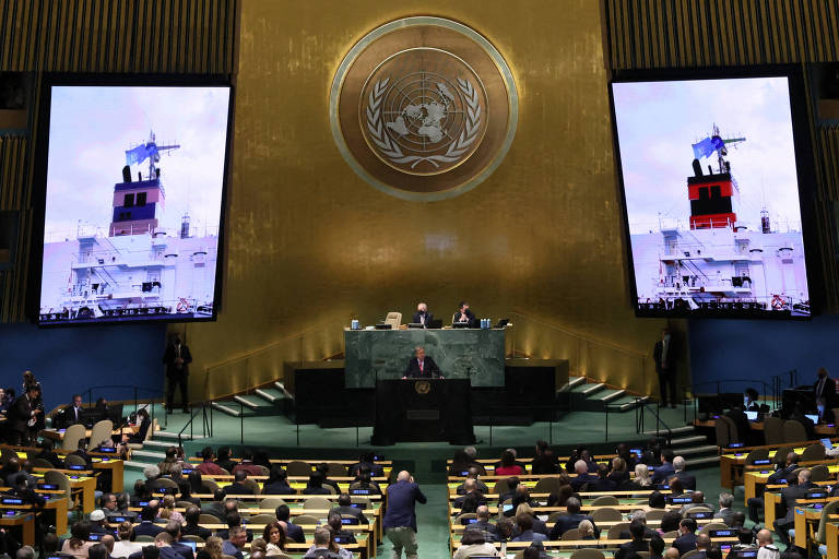 No salão da ONU, foto de navio cargueiro aparece em grandes telões enquanto Guterres discursa; a plateia está lotada