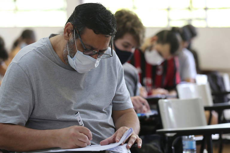 Estudantes fazem prova, um homem em primeiro plano usa máscara na sala de aula