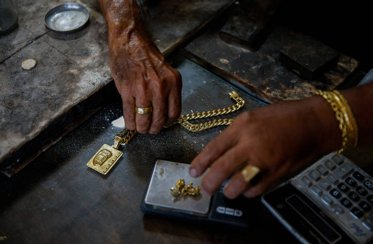 Mãos adornadas com anéis e pulseiras mexem em pepitas de ouro em cima de uma mesa; ao lado há um colar com um pingente grande de ouro e também uma calculadora