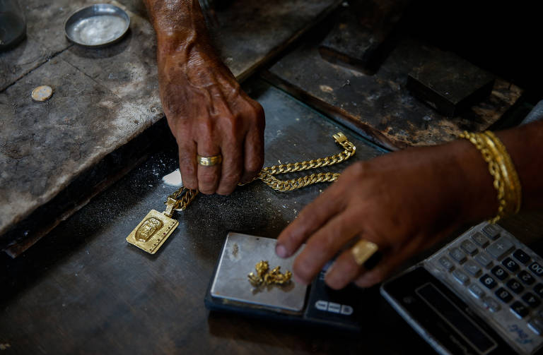 A imagem mostra duas mãos manipulando joias de ouro sobre uma balança. Uma corrente de ouro e um pingente estão sendo pesados. O fundo é uma superfície de trabalho escura, com um recipiente pequeno e uma calculadora visíveis.