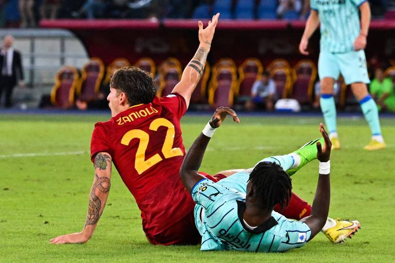 Sentado no campo, com o número 22 nas costas da camisa vermelha da Roma, Nicolo Zaniolo reclama de pênalti cometido por Caleb Okoli, do Atalanta, que está deitado a seu lado, em partida do Campeonato Italiano