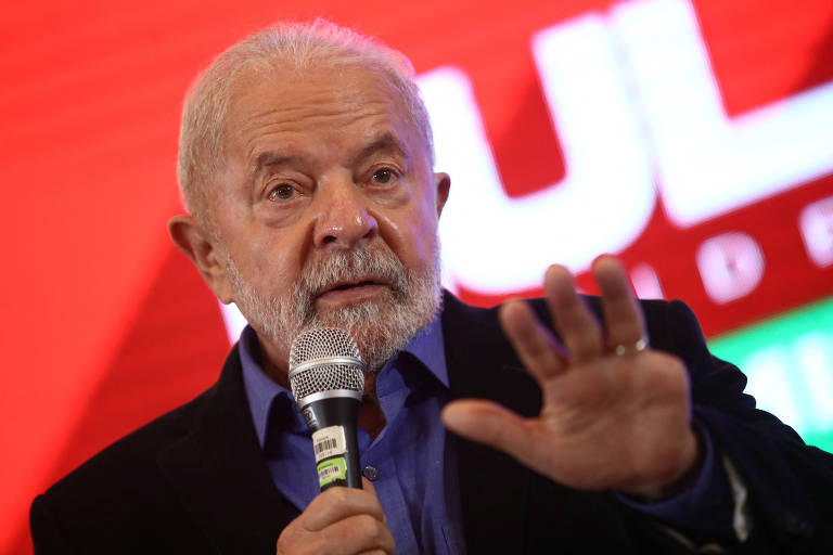Lula, homem branco de barba grisalha, segura microfone com a mão direita e estende a esquerda, que não tem o dedo mínimo. Fundo vermelho onde se lê as letras U e L do nome LULA em branco