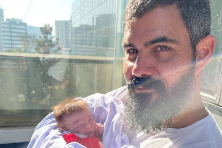 Homem branco com barba e cabelos castanho escuro segurando bebê recém-nascido em frente à janela 