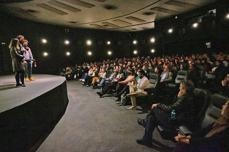 Foto interna de cinema lotado, pouco antes da exibição. No palco, três pessoas falam com o público