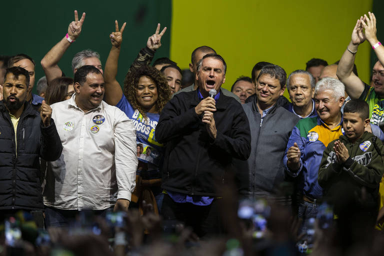 O presidente Jair Bolsonaro (PL) participa de comício nesta quarta-feira (28) no Centro de Convenções da Ponta da Praia, em Santos. Ele veste uma jaqueta preta e segura um microfone para discursar, ao lado de aliados políticos