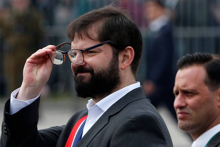 O presidente do Chile, Gabriel Boric, segura com a mão direita seus óculos, colocando-os no rosto, durante parada comemorativa de aniversário da independência do país; ele usa barba e bigode e veste um terno cinza, camisa branca e gravata nas cores azul e vermelha