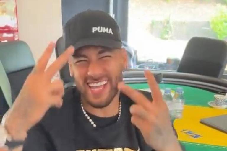 Neymar está de casaco preto com "Puma" em amarelo estampado no peito. Ele faz o número 2 com cada uma das mãos. Está de boné escuro