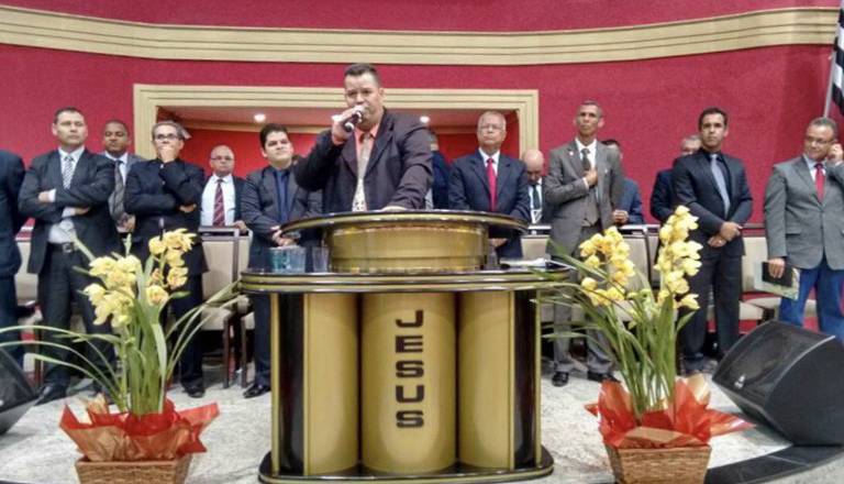 Foto colorida mostra Francys Lins discursando frente a um púlpito onde se lê a palavra 'Jesus'