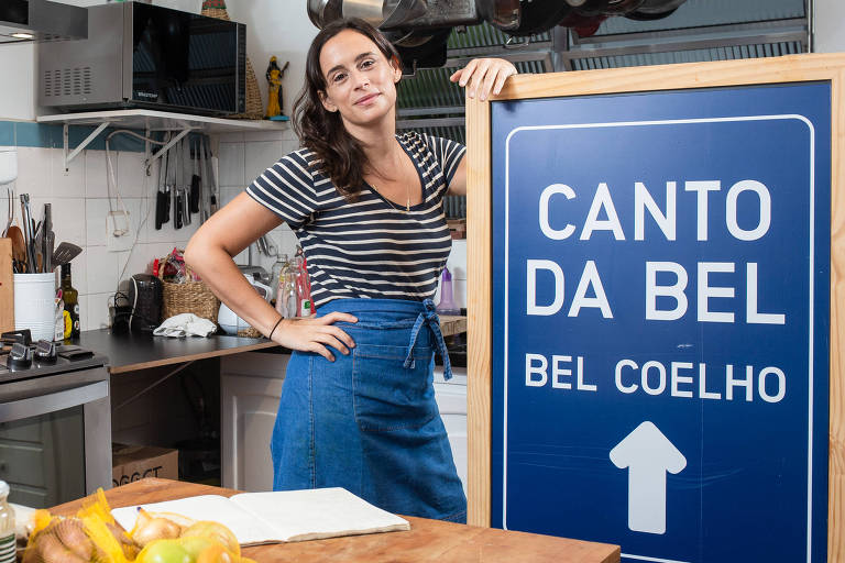 Chef Bel Coelho de blusa listrada e saia jeans em uma cozinha cheia de utensílios, apoiada em uma placa onde se lê "Canto da Bel"
