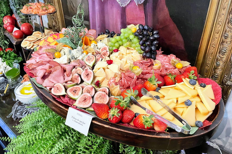A foto mostra uma travessa de madeira com muitos queijos, frios e frutas