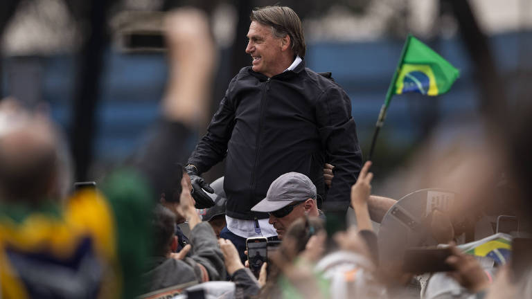 Em meio a apoiadores, alguns com bandeiras do Brasil, Bolsonaro está em pé, de blusa preta, olhando para o lado