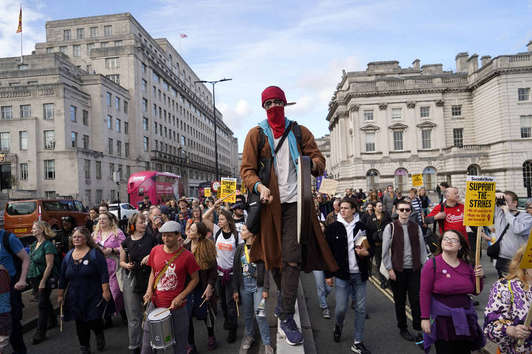 muitas pessoas em uma rua, com um dos manifestantes usando perna de pau e uma touca vermelha