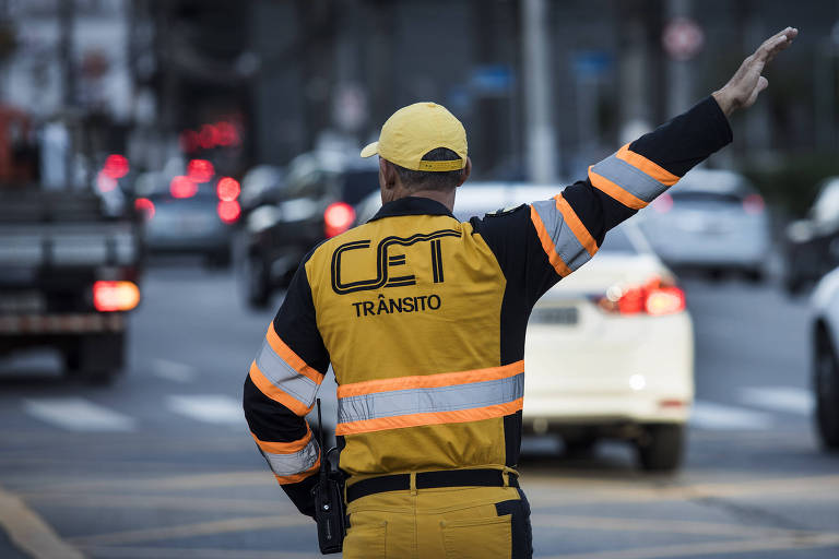 Agente da CET orienta trânsito no cruzamento das avenidas Faria Lima e Juscelino Kubitschek. Seu uniforme é amarelo, usa boné. Está de costas, é possível ver os carros ao fundo.