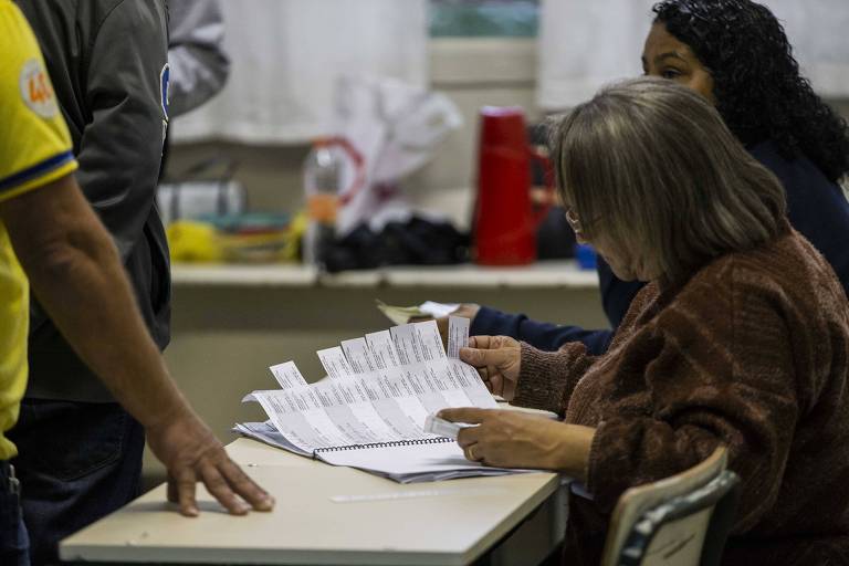 À esquerda, braço de pessoa de camiseta da seleção com mão apoiada em mesa branca e mesária com livro dos comprovantes de votação