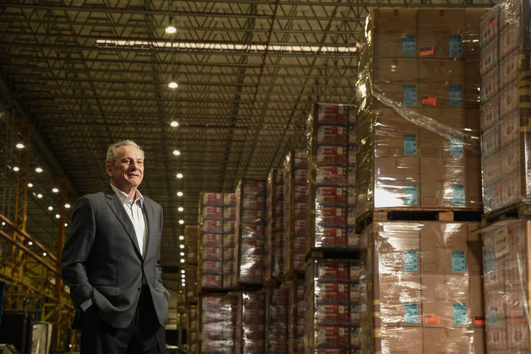 Homem branco de terno cinza, dentro de um centro de distribuição, com pilhas de caixas de papelão.