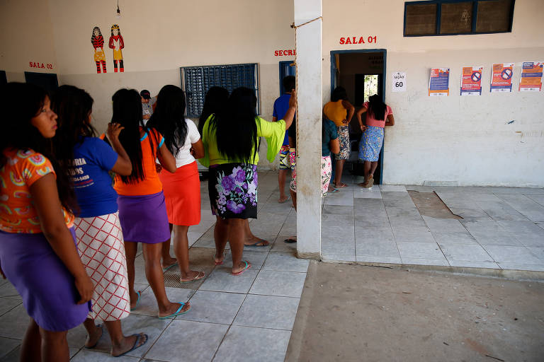 Fila de mulheres com roupas coloridas. a última da fila tem feição indígena. desenho de dois indígenas na parede branca