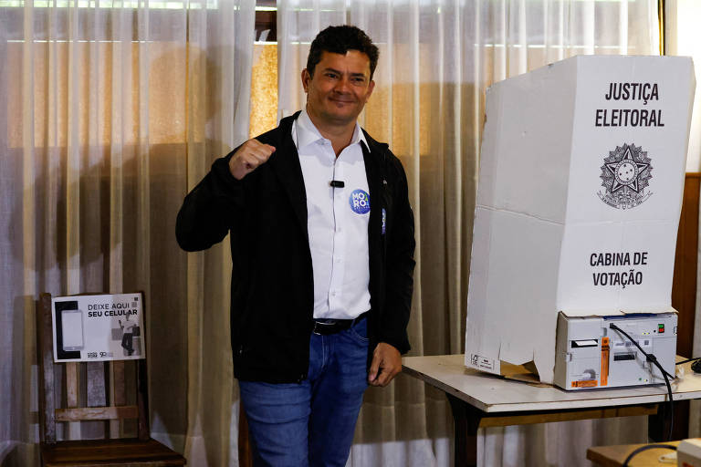 Ao lado de uma cabine de votação, um homem branco, de jeans, camisa branca e blusa preta, faz gesto com a mão fechada com uma das mãos. Moro é um homem branco, de cabelos escuros