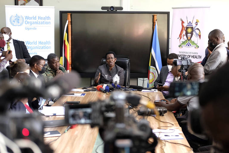A secretária do Ministério da Saúde em Uganda, Diana Atwine, uma mulher negra, de cabelos curtos e usando terno e óculos. Ela está sentada na ponta de uma mesa rodeada de jornalistas, com câmeras, celulares e microfones.