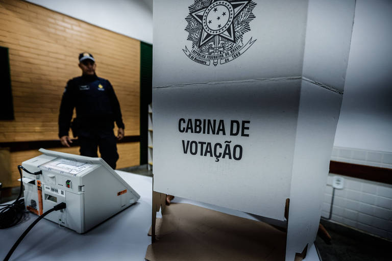 Cabine de votação e uma urna em primeiro plano e um policial ao fundo