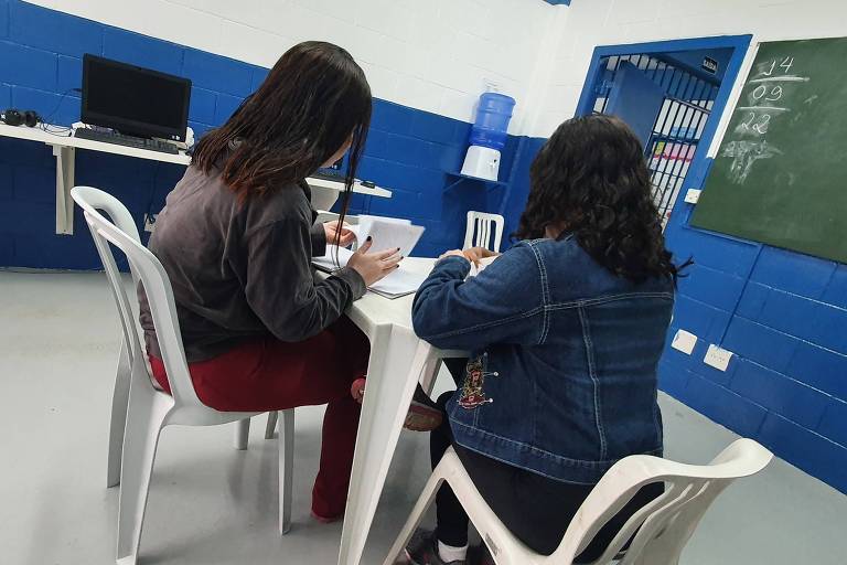 Duas meninas, sentadas ao redor de uma mesa de plástico, em sala de informática de centro da Fundação Casa. Elas estão de costas para a câmera.