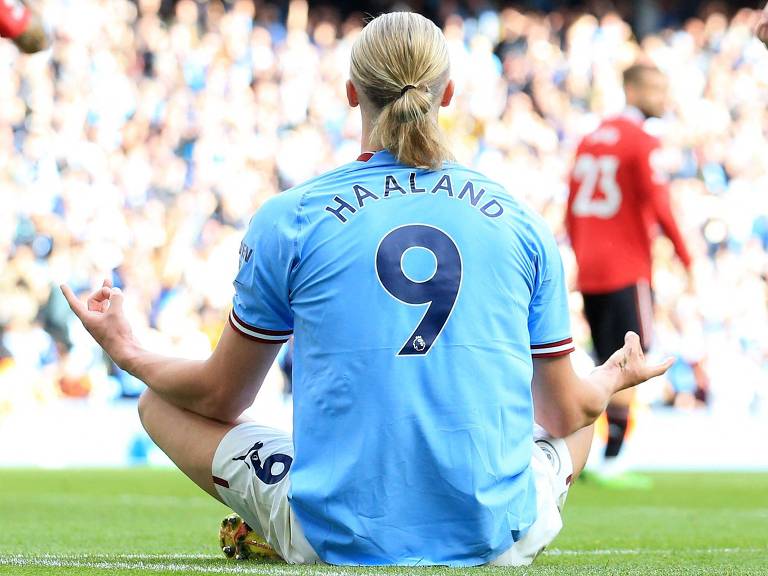 Sentado no gramado, número 9 na camisa azul celeste, Haaland faz posição de meditação, com o uso das duas mãos, para comemorar gol pelo Manchester City em clássico diante do Manchester United no Campeonato Inglês