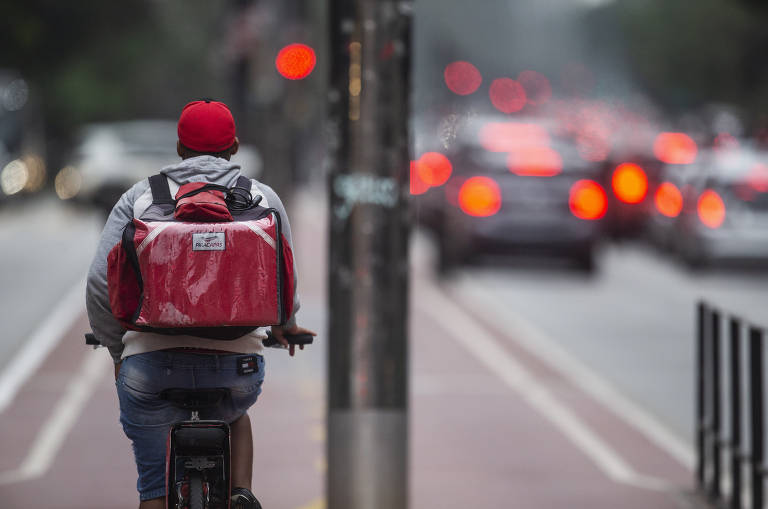 À direita da imagem, um entregador de costas pedala na ciclovia na avenida Paulista, em São Paulo. Ele usa uma bermuda jeans, um casaco cinza e um boné vermelho. Nas costas, carrega uma mochila de entrega vermelha