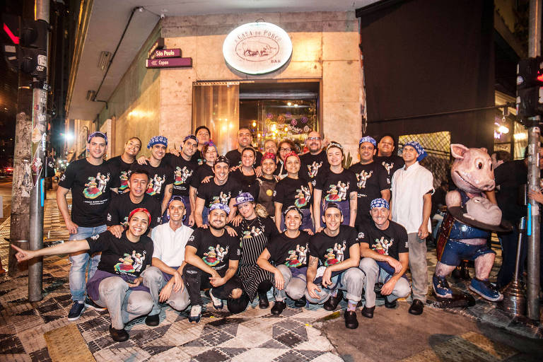 A foto mostra um grupo de pessoas vestindo camisetas pretas, em frente ao restaurante A Casa do Porco
