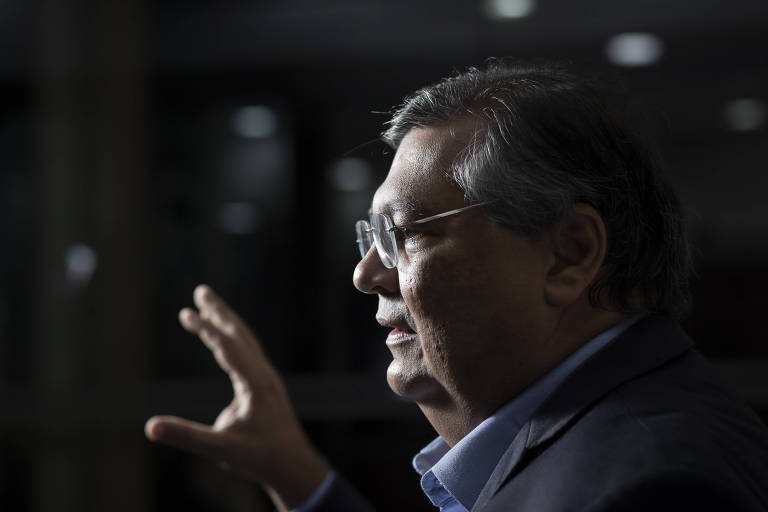 Foto mostra o senador eleito Flávio Dino (PSB-MA). Ele está de lado, em um fundo escuro. Ele veste terno, sem gravata e usa óculos. Gesticula com as mãos.
