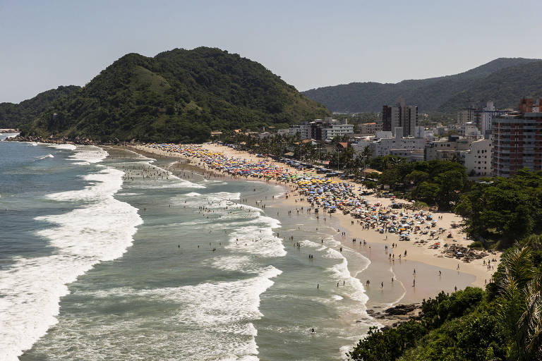 Imagem aérea da praia do Tombo, no Guarujá, com ondas grandes, muitas pessoas na praia e um morro ao fundo