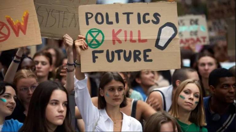 No meio de várias pessoas em um protesto contra a poluição do meio ambiente, mulher segura uma placa com a frase "Política mata o futuro"