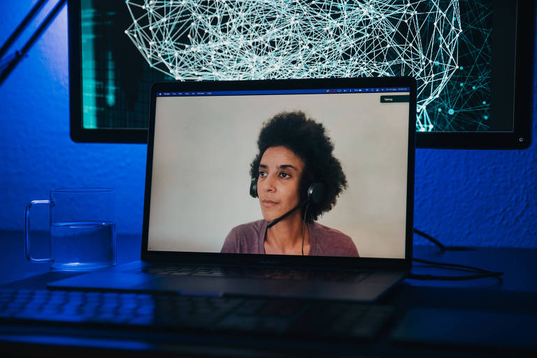 Fotografia colorida mostra um notebook em uma mesa; nele, está o retrato de uma mulher negra com cabelo crespo volumoso; ela usa uma camiseta e um fone de ouvido