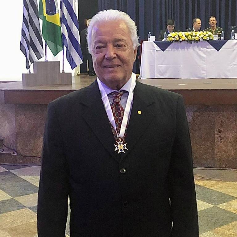Daniel Carlos dos Santos Ferreira, um homem branco, idoso e de cabelos brancos. Ele posa para a foto sorrindo, no Círculo Militar de SP, usando terno e uma medalha no pescoço.