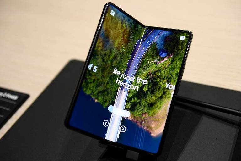 Smartphone Galaxy Z Fold 3 dobrável da Samsung; ele está sobre uma mesa e aparece em primeiro plano, com uma tela dupla, que exibe uma foto de satélite de mata verde e mar abaixo