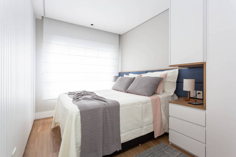 Quarto com cama de casal e mesa de cabeceira embutida no armário. O cômodo tem como cores predominantes o branco e o cinza claro