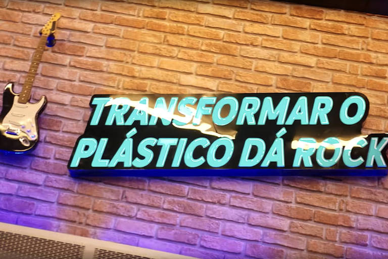 Reprodução colorida de vídeo com um letreiro azul claro pendurado em uma parede de tijolos: "Transformar o plástico dá rock", que aparece entre duas guitarras