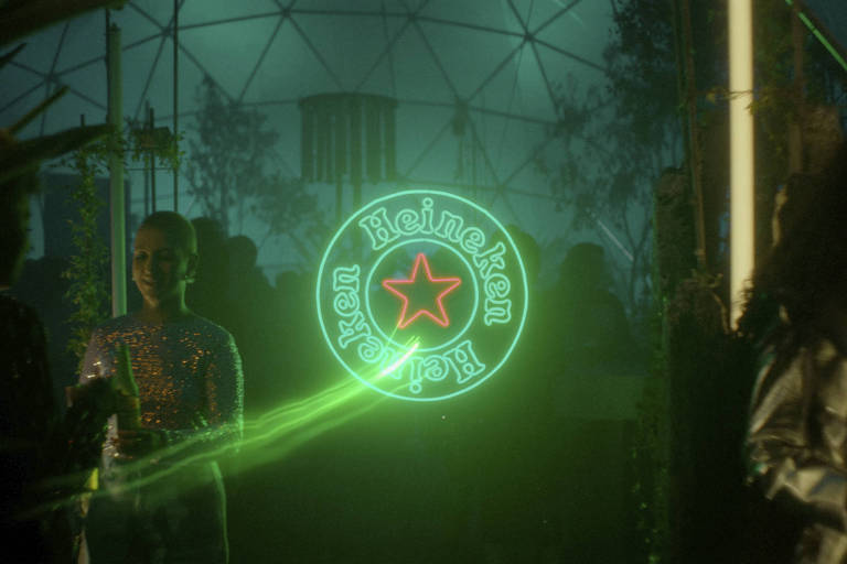 Fotografia com letreiro da Heineken em ambiente escuro com uma mulher à esquerda; luz verde prevalece