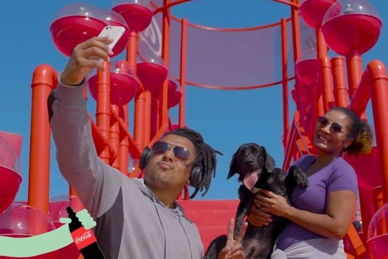Fotografia colorida mostra um homem negro à esquerda, tirando uma selfie, com uma mulher negra, que segura um cachorro preto; eles estão à ferente de uma estrutura vermelha em um ambiente azul em que se vê um dia ensolarado e de céu azul e uma