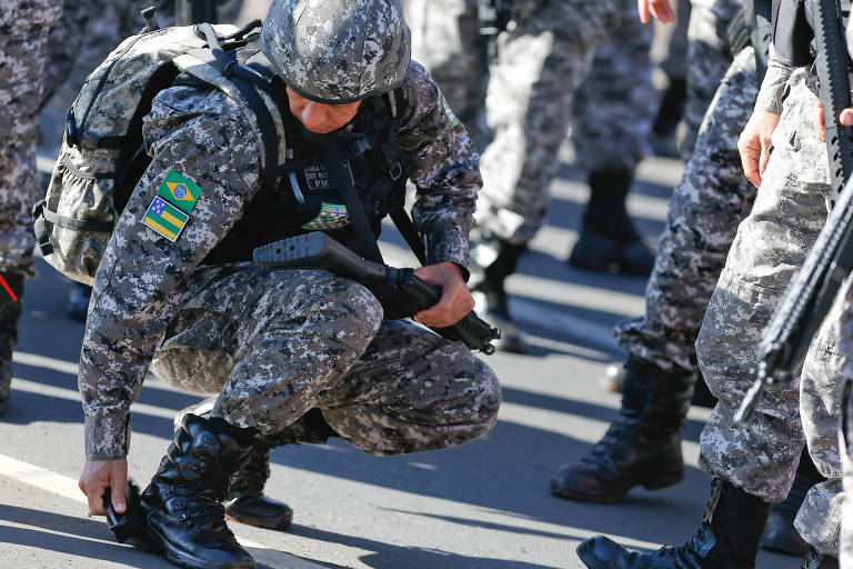 Soldado branco de capacete e roupa camuflada em tons de verde está agachado, aparentemente pegando algo no chão. Ao redor dele, soldados em pé