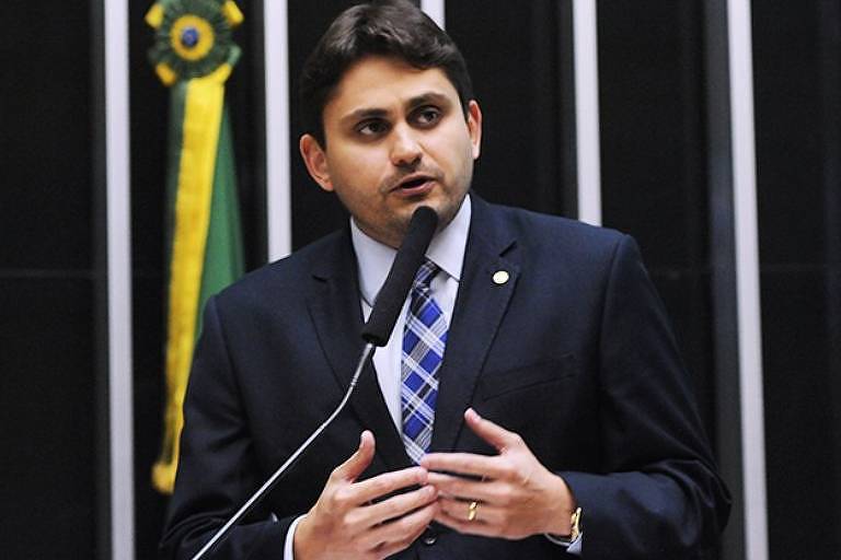 Juscelino Filho, um homem branco de cabelo preto curto, discursa diante do microfone. Ele tem as duas mãos em frente ao corpo